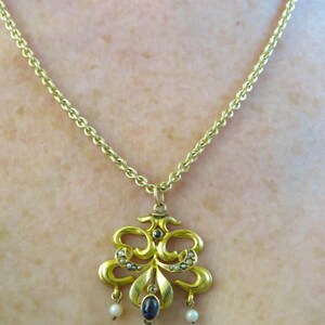 May include: A gold pendant necklace with an elaborate, symmetrical design. The pendant has a central dark blue stone, small pearls, and a fine chain. The necklace is likely vintage or antique.