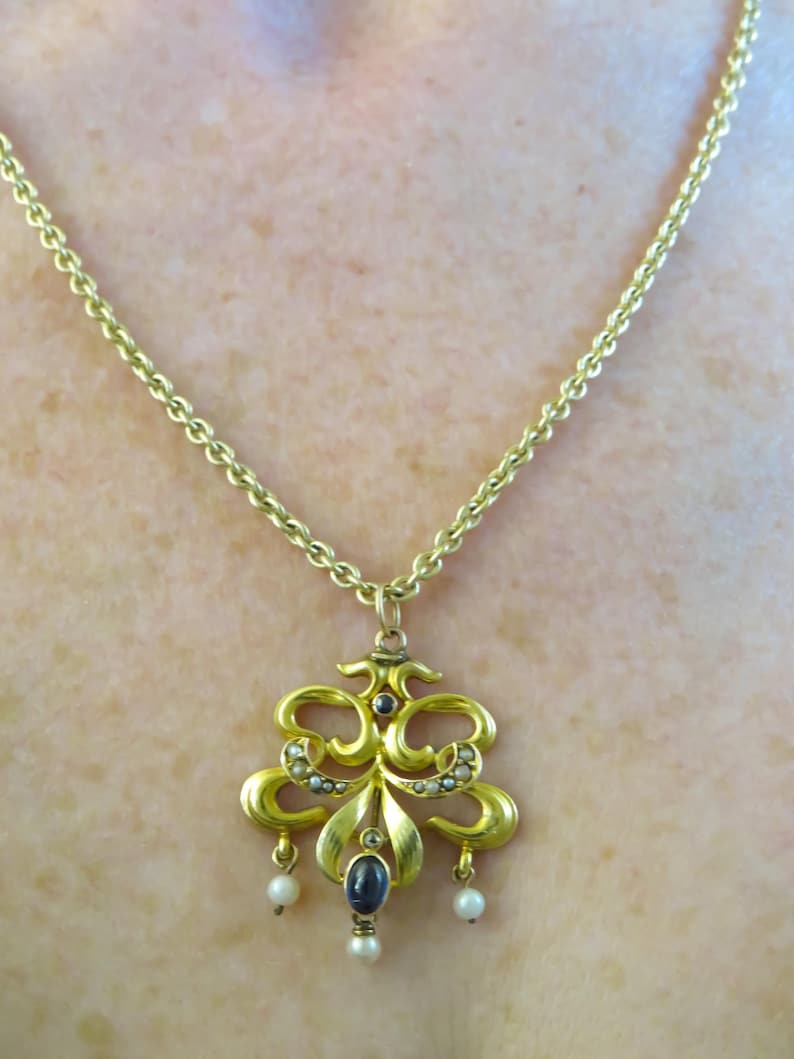May include: A gold pendant necklace with an elaborate, symmetrical design. The pendant has a central dark blue stone, small pearls, and a fine chain. The necklace is likely vintage or antique.