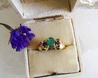 Könnte beinhalten: Ein goldener Ring mit einem ovalen Smaragd-Mittelstein und zwei kleineren dunkelblauen Steinen auf beiden Seiten. Der Ring wird in einer offenen, cremefarbenen Schmuckschatulle präsentiert. Ein Zweig lila Blumen liegt daneben.