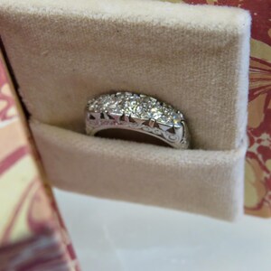 Op de afbeelding: Een zilveren ring met meerdere kleine diamanten in een rij, gepresenteerd in een beige fluwelen sieradendoosje. De ring heeft een sierlijk ontwerp op de band. De doos heeft een decoratief bloemenpatroon.