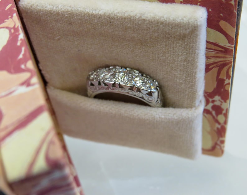 Op de afbeelding: Een zilveren ring met meerdere kleine diamanten in een rij, gepresenteerd in een beige fluwelen sieradendoosje. De ring heeft een sierlijk ontwerp op de band. De doos heeft een decoratief bloemenpatroon.