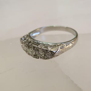 Op de afbeelding: Een zilveren ring met een rij van vijf heldere edelstenen. De ring heeft een sierlijk ontwerp met bloemen- en kruldetails op de band. De ring is geplaatst tegen een lichte achtergrond.