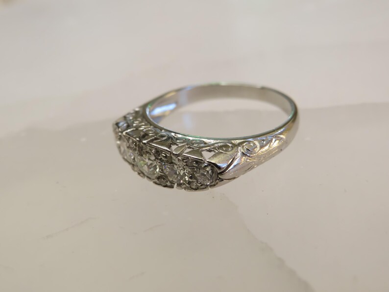Op de afbeelding: Een zilveren ring met een rij van vijf heldere edelstenen. De ring heeft een sierlijk ontwerp met bloemen- en kruldetails op de band. De ring is geplaatst tegen een lichte achtergrond.