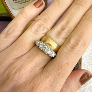 Op de afbeelding: Een gouden bandring en een platina ring met vijf diamanten worden getoond. De ringen zitten aan een hand, met een doos op de achtergrond. Op de doos staat "By Appointment Mappin & Webb Ltd Regent Street, London, W."
