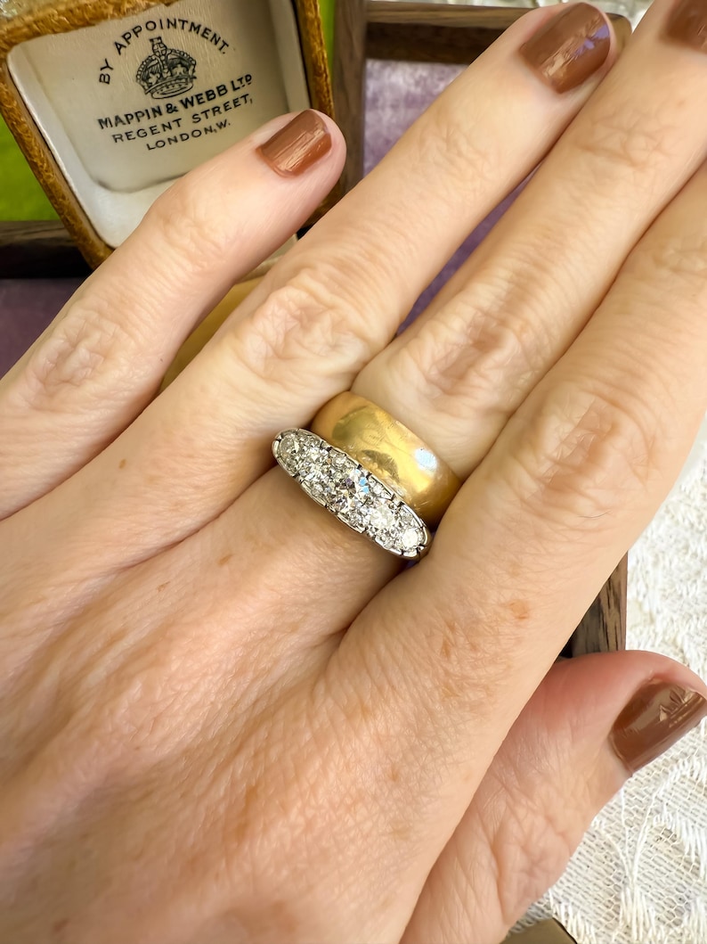 Op de afbeelding: Een gouden bandring en een platina ring met vijf diamanten worden getoond. De ringen zitten aan een hand, met een doos op de achtergrond. Op de doos staat "By Appointment Mappin & Webb Ltd Regent Street, London, W."