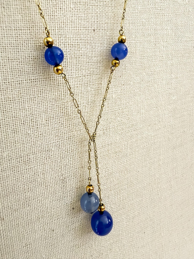 May include: A gold-toned necklace with a Y-shaped design. The necklace features three vibrant blue spherical beads and smaller gold-colored beads. The chain is delicate, with a simple link pattern. The necklace is displayed against a neutral background.