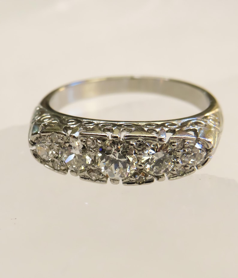 Op de afbeelding: Een zilveren ring met een rij van vijf heldere, rond geslepen edelstenen. De band heeft een sierlijk, gestructureerd ontwerp. De ring staat tegen een witte achtergrond, die de glans en ingewikkelde details benadrukt. Een klassiek sieraad.