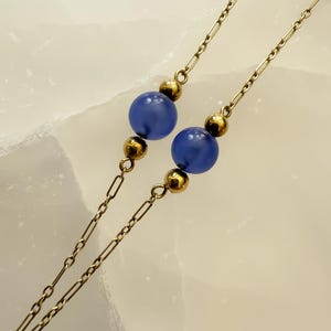 May include: Close-up of a necklace featuring two strands of delicate gold-colored chain. Each strand has a series of small, rectangular links and is punctuated by gold beads and two large, round, blue beads. The background is a light, textured surface.