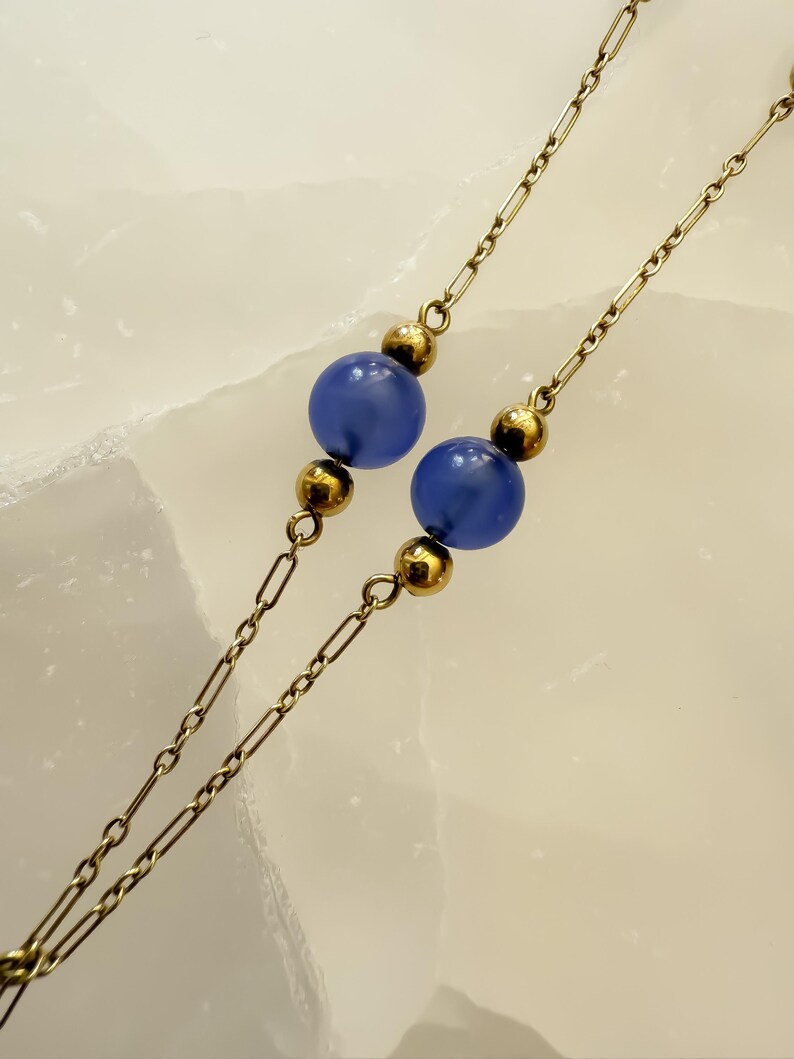 May include: Close-up of a necklace featuring two strands of delicate gold-colored chain. Each strand has a series of small, rectangular links and is punctuated by gold beads and two large, round, blue beads. The background is a light, textured surface.