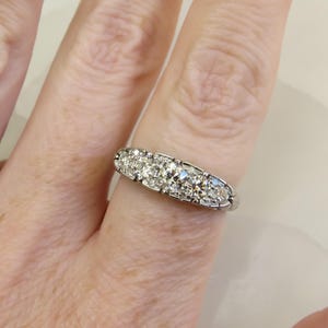 Op de afbeelding: Een zilveren ring met vijf heldere, ronde edelstenen. De ring wordt getoond op een vinger, met de hand en de achtergrond enigszins wazig. De edelstenen zijn in een rij gezet.