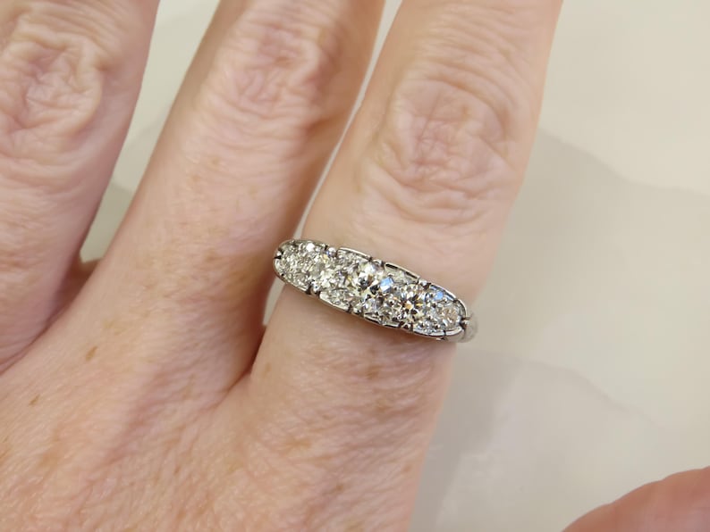 Op de afbeelding: Een zilveren ring met vijf heldere, ronde edelstenen. De ring wordt getoond op een vinger, met de hand en de achtergrond enigszins wazig. De edelstenen zijn in een rij gezet.