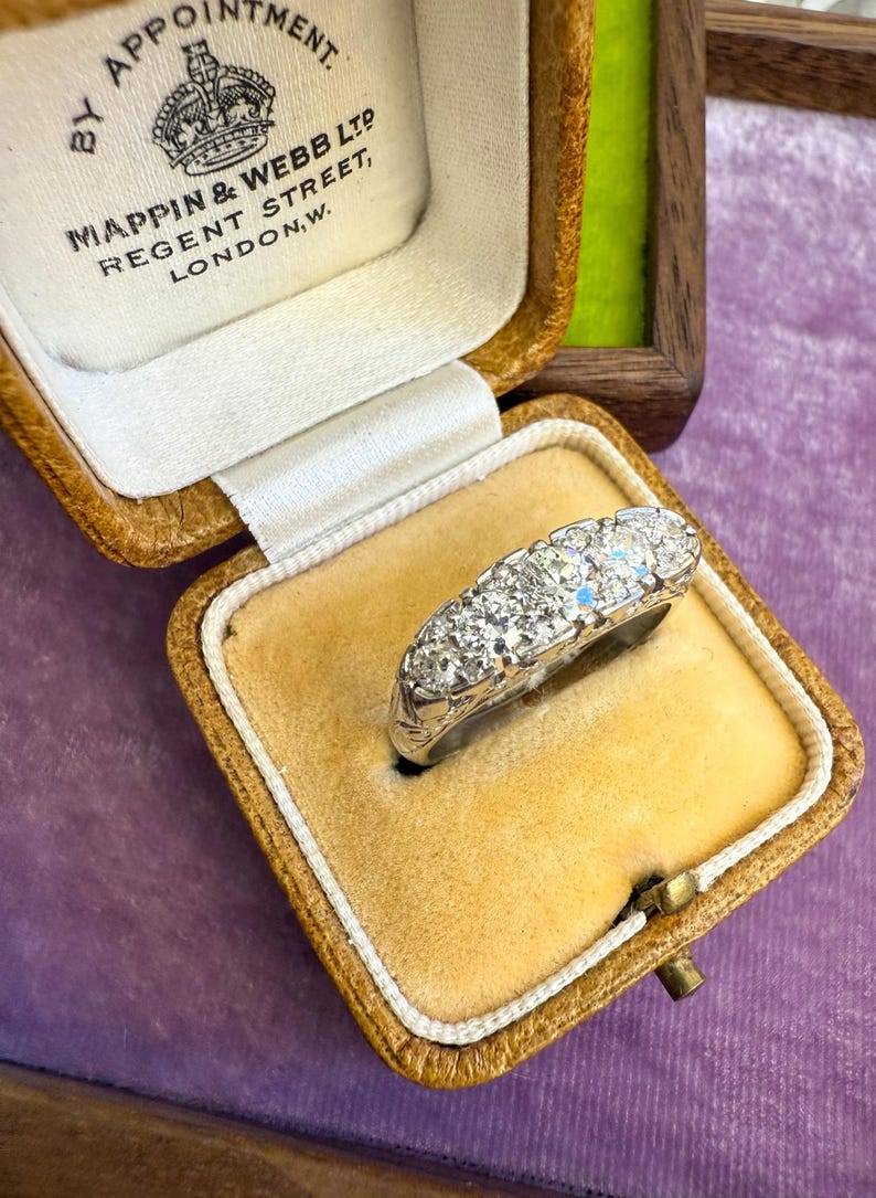 Op de afbeelding: Een vintage diamanten ring, gepresenteerd in de originele Mappin & Webb Ltd. doos. De ring heeft meerdere rond geslepen diamanten gezet in een witmetalen band. De doos is van bruin leer met een cr&egrave;mekleurig satijnen interieur.