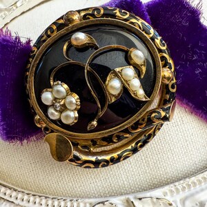 May include: An antique brooch with a black oval center, adorned with gold filigree and faux pearls. The brooch features a floral design with a cluster of pearls and a small diamond accent. The brooch is set against a purple velvet background.