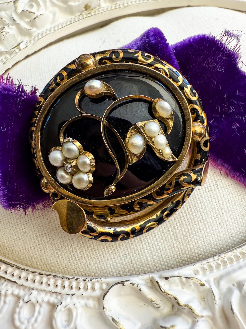May include: An antique brooch with a black oval center, adorned with gold filigree and faux pearls. The brooch features a floral design with a cluster of pearls and a small diamond accent. The brooch is set against a purple velvet background.