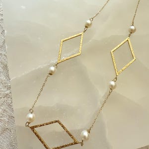 May include: A delicate gold-toned necklace featuring three diamond-shaped pendants with textured edges. The necklace is adorned with small, round pearls, creating an elegant and sophisticated design. The chain is fine and the necklace is displayed on a white surface.