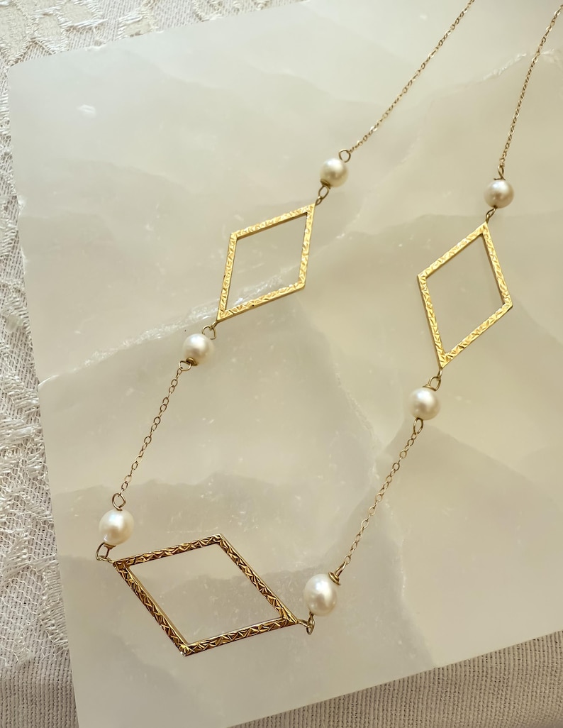 May include: A delicate gold-toned necklace featuring three diamond-shaped pendants with textured edges. The necklace is adorned with small, round pearls, creating an elegant and sophisticated design. The chain is fine and the necklace is displayed on a white surface.