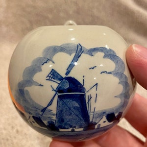 May include: A round, white ceramic ornament with a blue windmill design. The ornament features a traditional Dutch windmill scene with blue accents and a small loop for hanging. The design is hand-painted.