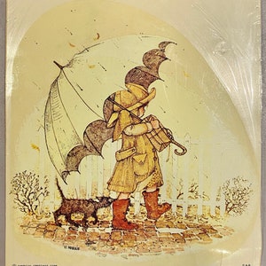 May include: A sheet of water transfer decals featuring a girl with a brown umbrella walking with a black cat. The girl is wearing a yellow coat, a brown hat, and red boots. The image is set against a white picket fence and a backdrop of a light brown background. The text "Holly Hobbie Decals" is printed at the top of the image.