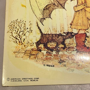 May include: A vintage illustration of a girl walking a black cat in the rain. The girl is wearing a brown coat and boots. The cat is walking in front of her, and there is a small tree in the background. The text "H. Hobbie" is signed in the bottom right corner. The text "AMERICAN GREETINGS CORP. CLEVELAND, U.S.A. MCM LXX" is in the bottom left corner.