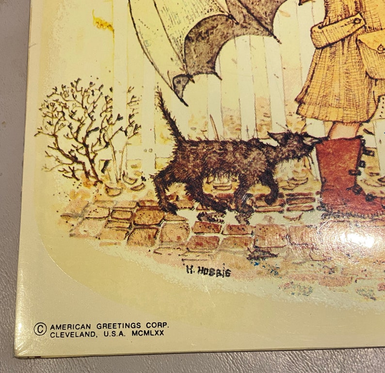 May include: A vintage illustration of a girl walking a black cat in the rain. The girl is wearing a brown coat and boots. The cat is walking in front of her, and there is a small tree in the background. The text "H. Hobbie" is signed in the bottom right corner. The text "AMERICAN GREETINGS CORP. CLEVELAND, U.S.A. MCM LXX" is in the bottom left corner.