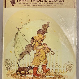 May include: A vintage Holly Hobbie decal sheet featuring a girl walking with a black cat under an umbrella. The decal sheet is suitable for decoupage craft use and interior decorating. It measures 8 inches by 9 inches.