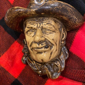 May include: A ceramic wall sculpture of a cowboy wearing a brown hat and a brown jacket. The cowboy has a mustache and is holding a cigar in his mouth.