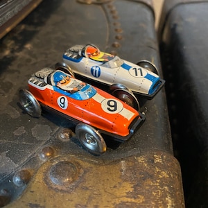 May include: Two vintage toy race cars, one orange and black with the number 9, the other white with the number 11, on a textured brown surface.