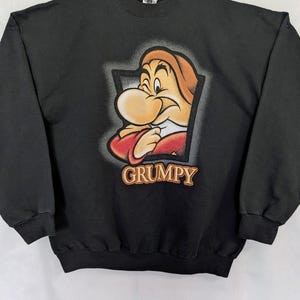Peut inclure: Sweat-shirt noir avec un dessin de Grincheux de Blanche-Neige. Le personnage de dessin animé a un grand nez, des cheveux orange et une chemise rouge. Le mot "GRUMPY" est imprimé en lettres dorées sous le personnage.