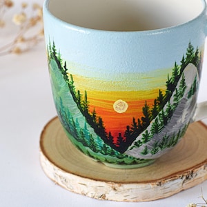 Può includere: Una tazza in ceramica bianca con una scena dipinta a mano di una catena montuosa al tramonto. La tazza ha un cielo azzurro chiaro, un tramonto arancione e giallo e alberi verdi. La tazza è appoggiata su un sottobicchiere di legno.