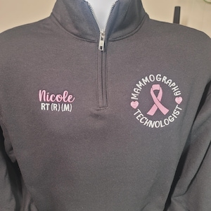May include: Black quarter-zip sweatshirt with a silver zipper. The left chest has the name "Nicole" embroidered in pink, with "RT (R) (M)" below. The right chest features a pink ribbon and the words "Mammography Technologist" in a circle.