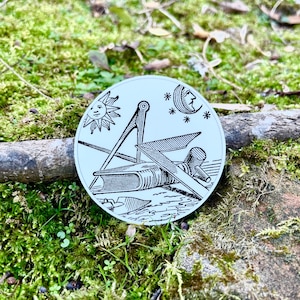 May include: A white circular sticker with a black and white illustration of a compass, sun, moon, and stars. The compass is pointing north.