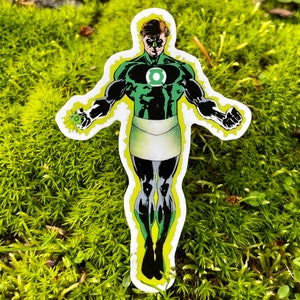 May include: Green Lantern comic book character sticker with a green and black costume, a white belt, and a green glowing aura. The sticker is on a green mossy surface.