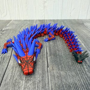 May include: A red and blue articulated dragon toy with a spiderweb design on the head. The dragon has blue spikes along its back and a black head and tail. The toy is made of plastic and is on a wooden surface.