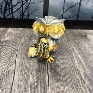 May include: A metallic owl figurine with gold and silver accents. The owl has large, round eyes and detailed feather-like structures. The figurine is posed on a wooden surface, with a window and bare tree branches in the background. The owl's design suggests a steampunk or mechanical theme.