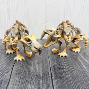 May include: Two beige and tan saber-toothed tiger skeleton models. The models are posed facing each other, with their large teeth visible. The skeletons are on a gray wooden surface.