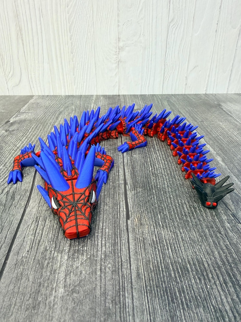 May include: A red and blue articulated dragon toy with a spiderweb design on the head. The dragon has blue spikes along its back and a black head and tail. The toy is made of plastic and is on a wooden surface.