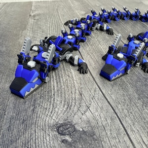May include: A blue and black articulated toy dragon with silver accents. The dragon is made of multiple connected segments, allowing for flexible posing. The head features a detailed design with sharp teeth. The toy is designed to resemble a robotic dragon.