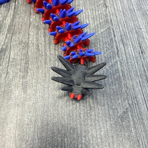 May include: A segmented, articulated sculpture with a red and blue color scheme. The main body is red with blue spikes. A black, spidery-looking head with red accents is at the end. The sculpture is on a gray wooden surface.
