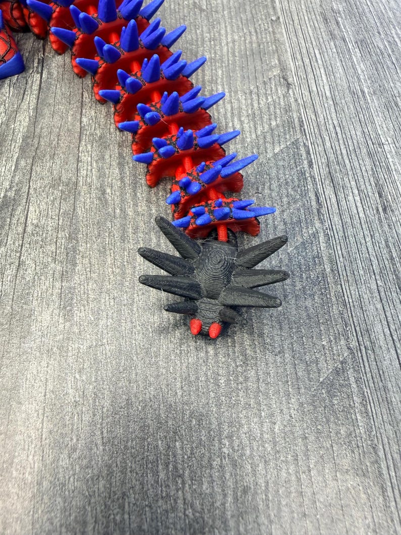 May include: A segmented, articulated sculpture with a red and blue color scheme. The main body is red with blue spikes. A black, spidery-looking head with red accents is at the end. The sculpture is on a gray wooden surface.