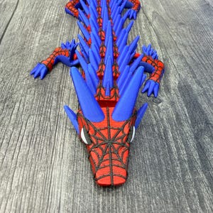 May include: A 3D-printed dragon toy in blue and red. The dragon has a spiky spine, webbed face, and articulated limbs. The toy is lying on a wooden surface. The dragon's head features a spiderweb design.
