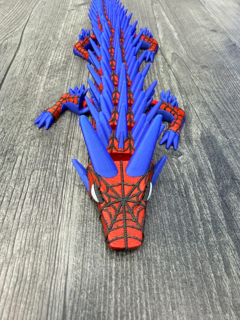 May include: A 3D-printed dragon toy in blue and red. The dragon has a spiky spine, webbed face, and articulated limbs. The toy is lying on a wooden surface. The dragon's head features a spiderweb design.