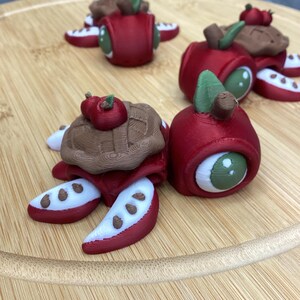 May include: A collection of whimsical, 3D-printed apple-shaped creatures with brown tops, green eyes, and red and white accents. The creatures are arranged on a wooden surface, showcasing their unique design and playful aesthetic.
