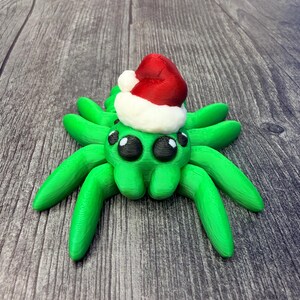 May include: A green spider figurine with eight legs and two large black eyes. The spider is wearing a small red and white Santa hat. The figurine is on a wooden surface.