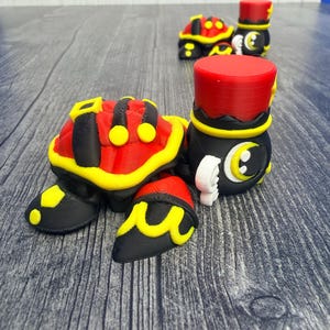 May include: 3D-printed turtle figurines in black, red, and yellow. The turtle shells are red with yellow accents, and the feet are black with yellow details. One figurine is in the foreground, with two more in the background. The figurines have a whimsical, cartoonish design.