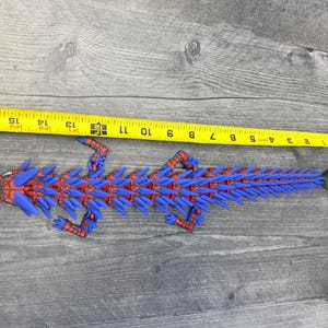 May include: A red and blue articulated lizard toy, approximately 12 inches long, with a spiky design. The toy is measured with a yellow tape measure on a wooden surface.