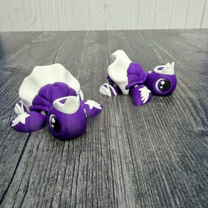 May include: Two 3D-printed turtle figurines. The turtles are primarily purple with white accents on their shells, feet, and heads. They have large, round eyes and are lying on a gray wooden surface. The turtles appear to be decorative items.