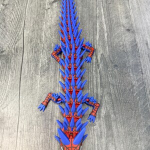 May include: A 3D-printed, stylized alligator figurine in red and blue. The alligator has a red body with blue spikes along its back and head. The head features a spiderweb design. The figurine is lying on a wooden surface.
