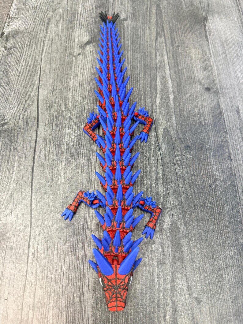 May include: A 3D-printed, stylized alligator figurine in red and blue. The alligator has a red body with blue spikes along its back and head. The head features a spiderweb design. The figurine is lying on a wooden surface.
