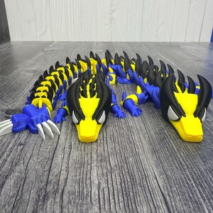 May include: A 3D-printed articulated dragon toy in blue, yellow, black, and white. The dragon has a segmented body, sharp claws, and a detailed head with expressive eyes. The toy is designed to be flexible and poseable.