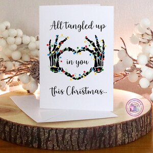 K&ouml;nnte beinhalten: Eine wei&szlig;e Gru&szlig;karte mit schwarzen Skeletth&auml;nden, die ein Herz bilden und mit bunten Lichterketten umwickelt sind. Der Text lautet: All tangled up in you this Christmas. Die Karte wird zusammen mit einem wei&szlig;en Umschlag pr&auml;sentiert.